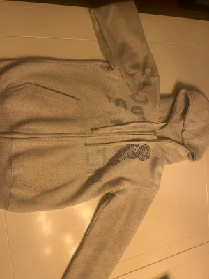 Grå hoodie med bling och tryck från von studio  - Säljer en ljusgrå hoodie med dragkedja och huva. På framsidan finns ett stort tryck i silverglittrande stenar med text och en tecknad figur. Hoodien har fickor på sidorna och ribbade muddar vid ärmar och nederkant. Perfekt för en chill och trendig look. Från von studio 
