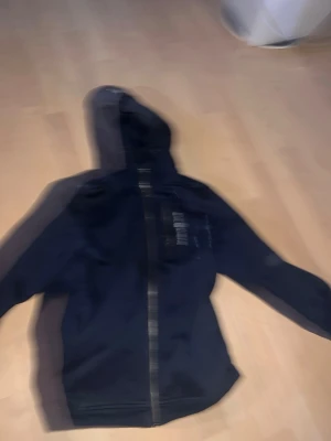 Mörkblå sailracing  - Säljer en svart hoodie med dragkedja framtill och huva. Hoodien har en ficka på bröstet och ribbade muddar vid ärmslut och nederkant. Perfekt för en avslappnad och chill stil.