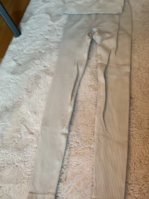 Beige ribbade leggings med hög midja - Säljer ett par beige leggings med ribbad struktur och hög midja. De är tighta och stretchiga, perfekta för en avslappnad och trendig look. Materialet känns mjukt och följsamt mot huden. Passar perfekt till både oversized tröjor och crop tops.