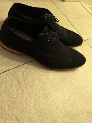 Svarta mockaloafers från Hugo - Stilrena svarta loafers från Hugo i mocka med klassisk snörning och låg klack. Skorna har en elegant, smal siluett och kontrasterande ljusbrun sula. Perfekta för dig som vill ha en dressad men avslappnad look. Ordinarie pris 3000 kr