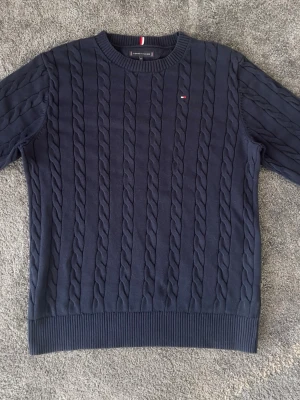 Mörkblå kabelstickad tröja Tommy Hilfiger - Snygg mörkblå kabelstickad tröja från Tommy Hilfiger med rund halsringning och diskret logga på bröstet. Tröjan har ribbade muddar vid ärmslut och nederkant, perfekt för en klassisk och stilren look. Knappt använd, nyskick. Storlek 164, passar dig runt 170cm.