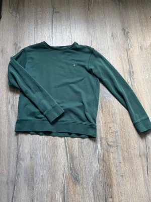 Mörkgrön sweatshirt från Gant - Mörkgrön sweatshirt från Gant med rund halsringning och diskret logga broderad på bröstet. Tröjan har långa ärmar och breda muddar vid ärmslut och nederkant. Perfekt för en avslappnad och stilren look.