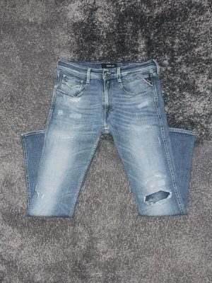 Blå slitna jeans från Replay - Säljer ett par ljusblå jeans från Replay med slitningar och hål på ena benet. Klassisk femficksmodell med skinny fit och dragkedjegylf. Snygg tvättad look och detaljer som ger en trendig vibe.