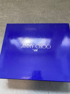Svarta pumps från Jimmy Choo x H&M st 37 - Snygga svarta pumps med hög klack från Jimmy Choo x H&M. Skorna har öppen tå, breda remmar med nitar och ögon, samt justerbart spänne runt ankeln. Tillverkade i läder med mycket fina detaljer. Aldrig använda. Alla tags plus låda och dustbag finns kvar. 