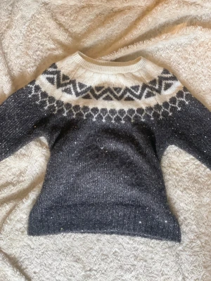 Mörkgrå stickad tröja GapKids XL - Mysig mörkgrå stickad tröja från GapKids med vitt och svart mönster runt halsen. Tröjan har rund halsringning och långa ärmar, perfekt för kalla dagar. Materialet är mjukt och glittrigt, vilket ger en extra touch till looken.