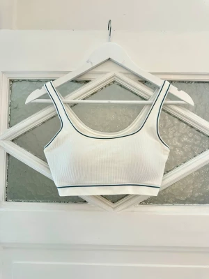 Vit bh/sporttopp med blå kant - Snygg vit ribbad body i crop-modell med breda axelband och blå kontrastkant längs ringningen och nederkanten. Tillverkad i mjukt, stretchigt material som sitter skönt mot kroppen. Perfekt att styla under skjortor eller tröjor.