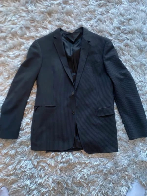 Svart kavaj från tiger of sweden - Snygg svart kavaj med tunna, diskreta ränder och två knappar framtill. Klassisk passform med två fickor och bröstficka. Fodrad insida och smal krage ger en stilren look. Perfekt för dig som vill ha en enkel men elegant kavaj.