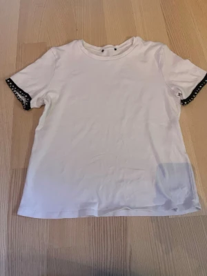 Vit t-shirt med pärldetaljer - Vit t-shirt med korta ärmar och svarta kanter dekorerade med pärlor. T-shirten har en klassisk rund halsringning och är gjord i mjukt bomullstyg. Perfekt för dig som vill ha en enkel men ändå unik look.