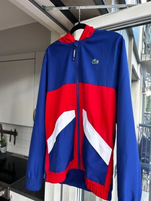 Blå och röd hoodie från Lacoste - Snygg hoodie från Lacoste i blått och rött med vita detaljer. Dragkedja framtill, huva med röd insida och klassisk Lacoste-logga på bröstet. Tillverkad i ett lätt material, perfekt för en sportig och avslappnad stil.
