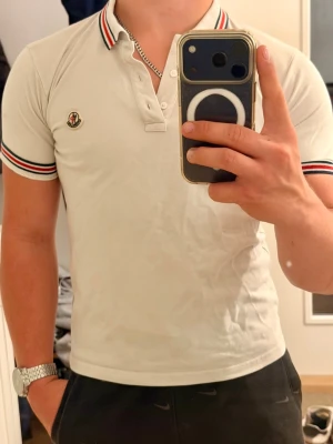 Vit Moncler pikétröja - Vit pikéskjorta från Moncler med klassisk krage och knappar framtill, tröjan är i storlek 164cm vilket motsvarar ungefär xs, den har en fläck vid nedre delen därav priset, skriv gärna för frågor eller funderingar!