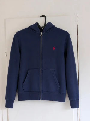Ralph Lauren zip hoodie - Polo Ralph Lauren zip-up hoodie i klassisk marinblå färg med röd broderad logga på bröstet 💙❤️ Storlek: M (10–12 år) • Hel dragkedja framtill • Huva • Två fickor framtill • Mjuk och varm insida • Ribbad mudd vid ärmar och nederkant • Klassisk Polo Ralph Lauren-design Fint begagnat skick utan defekter! Perfekt vardagshoodie som passar till det mesta ✨