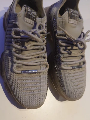Beige sneakers med strassdetaljer - Snygga beige sneakers med stickad ovandel och glittriga strassdetaljer på sidorna. Skorna har tjock, transparent sula och breda snören för en modern look. Perfekta för dig som vill sticka ut lite extra i vardagen.