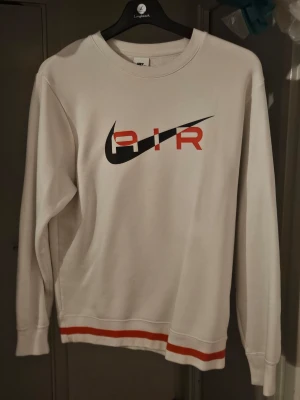 Vit Nike Air stickad tröja - Vit stickad tröja från Nike med AIR-tryck och svart swoosh på bröstet. Tröjan har långa ärmar och röda ränder vid nederkanten. Perfekt för dig som gillar sportig och clean stil.