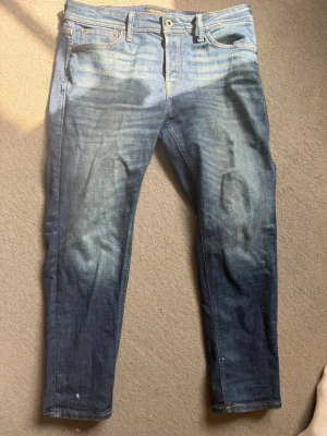 Blå taperd jeans från Jack & Jones - Snygga blå slim fit jeans från Jack & Jones med klassisk femficksdesign och slitna detaljer. Jeansen har en skön stretchig denimkvalitet och läderpatch bak i midjan. Perfekta till en avslappnad streetstyle-look. Storlek 34/30