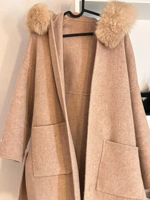 Beige kappa med äktapälsluva - Snygg beige kappa med stor luva dekorerad med fluffig äktapäls. Kappan har två stora fickor framtill och ett matchande knytbälte i midjan. Tillverkad i mjukt ull-liknande material och har en avslappnad, oversized passform.