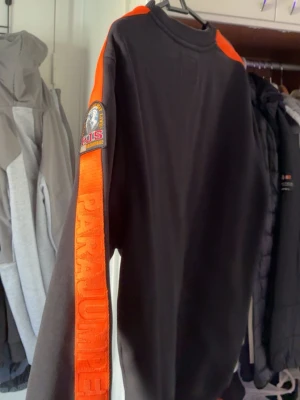 Parajumper  - Cool svart långärmad tröja från Parajumpers med orange ränder längs ärmarna och ett stort broderat märke på axeln. Tröjan har en sportig look och texten 'PARAJUMPERS' på ärmen. Perfekt för dig som gillar streetwear och snygga detaljer.