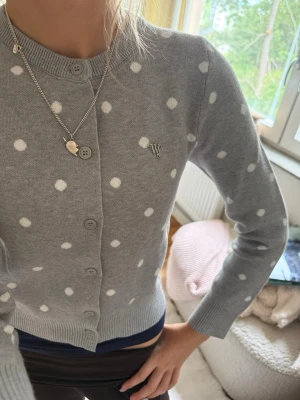 Grå prickig kofta från WA YA xxs  - Supersöt grå kofta från WA YA Collection med vita prickar och rund hals. Knäpps med matchande knappar framtill och har en liten metallogo på bröstet. Perfekt till jeans eller kjol för en lekfull look.
