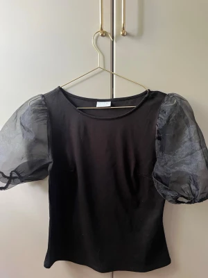 Svart topp med puffärmar  - Svart topp från Vila med transparenta puffärmar. Kroppsdelen är i mjukt tyg och toppen har en rund halsringning. 