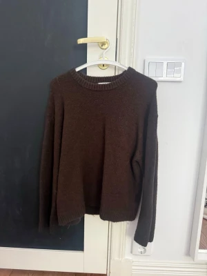 Brun stickad tröja med rund hals - Mysig brun stickad tröja med rund halsringning och långa ärmar. Tröjan har en enkel, rak passform och ribbstickade muddar vid hals, ärmslut och nederkant. Perfekt för kyliga dagar och enkel att matcha med olika outfits.