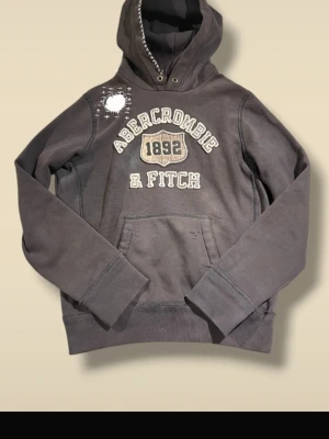 Mörkbrun hoodie Abercrombie & Fitch - Säljer en mörkbrun hoodie från Abercrombie & Fitch med broderad logga och texten '1892' på bröstet. Hoodien har en stor ficka framtill, huva med snörning och dekorativa sömmar samt ett vitt broderat märke på axeln. Perfekt för en avslappnad stil.