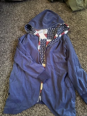 Blå hoodiejacka från L.B.C. med stjärnmönster - Blå hoodiejacka från L.B.C. med dragkedja och huva. Insidan av huvan och öppningen har ett mönster med vita stjärnor och röda ränder, inspirerat av amerikanska flaggan. Jackan har långa ärmar och ribbade muddar.