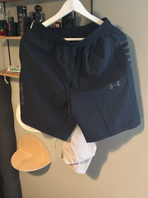 Under armour shorts  - Snygga svarta träningsshorts från Under Armour med elastisk midja och snörning. Shortsen har diskreta loggor på sidan och är tillverkade i ett lätt och snabbtorkande syntetmaterial. Perfekta för gymmet eller löprundan.
