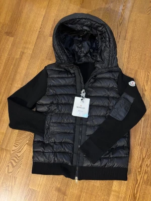 Moncler cardigan  - Säljer en svart pufferjacka från Moncler med stickade ärmar och huva. Jackan har dragkedja framtill, fickor och Moncler-logga på ärmen. Kroppen är quiltad och glansig medan ärmarna är ribbstickade för en snygg kontrast. Perfekt för kalla vinterdagar.