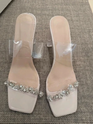 Beiga klackar med strass - Säljer ett par trendiga beiga klackar med transparenta remmar och fyrkantig tå. Skorna har klack i akryl och dekoreras med glittrande strass framtill. Perfekta för dig som vill ha en modern och elegant look. Det är storlek 40 men passar mer 39 