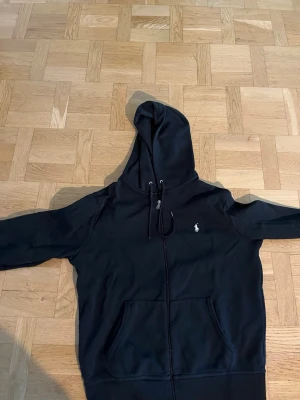 Ralph lauren hoodie - Säljer en svart hoodie med dragkedja och huva. Klassisk design med fickor framtill och justerbara snören i huvan. Liten vit logga broderad på bröstet. Perfekt för en avslappnad och stilren look.