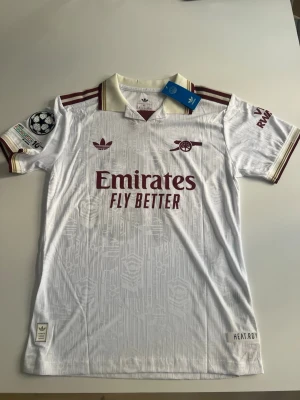 Helt ny! Arsenal 25/26 tredje Eze - Snygg vit Arsenal matchtröja från Adidas med vinröda detaljer och klubbmärke på bröstet. Tröjan har korta ärmar, krage och tryckta sponsorer samt Champions League-märke på ärmen. Namnet 'EZE' och nummer 10 på ryggen. Perfekt för fotbollsfans!