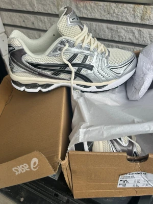 Asics Gel-Kayano 14 silver/vit - Säljer ett par Asics Gel-Kayano 14 sneakers i silver och vitt med svarta detaljer. Skorna har mesh- och syntetmaterial, chunky sula och klassisk Asics-logga på sidan. Perfekta för dig som gillar retrostil och sportig vibe.