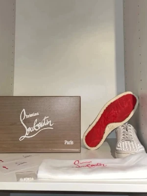 Vita Christian Louboutin sneakers med nitar - Unika vita sneakers från Christian Louboutin med ikoniska röda sulor och vita nitar över tån. Skorna har lågt skaft, snörning och är tillverkade i läder. Perfekta för dig som vill sticka ut med exklusiv design och edgy detaljer.