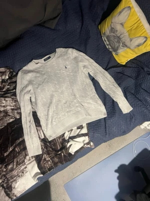 Ljusgrå kabelstickad tröja - Snygg ljusgrå kabelstickad tröja med rund hals och långa ärmar. Tröjan har ett diskret broderat emblem på bröstet och ribbade muddar vid ärmslut och nederkant. Perfekt för lager på lager och enkel att matcha med olika outfits.