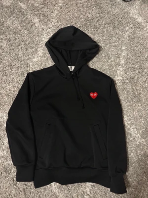 Svart hoodie med rött hjärta - Säljer en svart hoodie med huva och dragsko. På bröstet finns ett broderat rött hjärta med ögon, vilket ger en unik och trendig look. Hoodien har magficka och ribbade muddar vid ärmar och nederkant. Perfekt för dig som gillar grisch och vill sticka ut. Den är storlek S men passar bra på XS. Pris kan förhandlas
