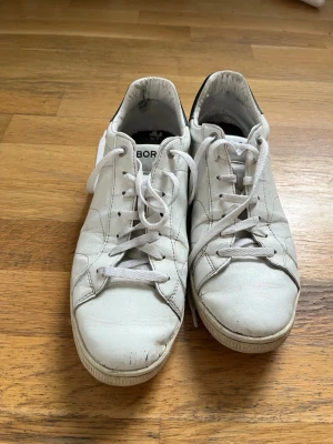 Vita Björn Borg sneakers i läder - Klassiska vita sneakers från Björn Borg i läder med svart detalj på hälen. Skorna har lågt skaft, vita snören och diskret logga på sidan. Perfekta för en clean och stilren look.