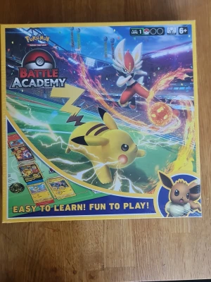 Pokémon Battle Academy brädspel sealed - Pokémon Battle Academy är ett färgglatt brädspel med Pikachu, Cinderace och Eevee på framsidan. Innehåller tre kompletta Pokémon TCG-lekar, spelbräde, instruktioner och tillbehör. Perfekt för två spelare som vill lära sig Pokémonkortspelet på ett enkelt och roligt sätt.