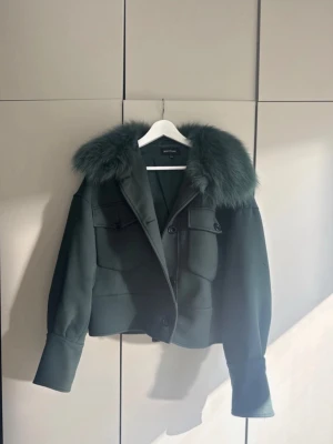 Mörkgrön pälsjacka från Meotine - Snygg mörkgrön pälsjacka från Meotine med stor fluffig fuskpälskrage. Jackan har två stora bröstfickor med lock och knappar, samt breda muddar vid ärmsluten. Kortare modell som ger en trendig siluett. Storlek S/M.