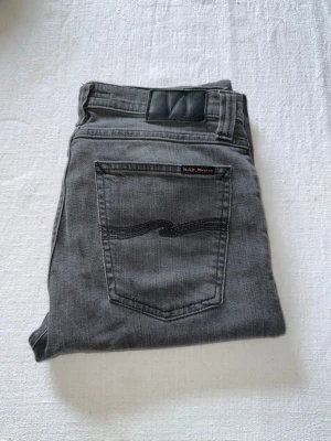 Gråa Nudie Jeans - Snygga gråa slim fit jeans med klassisk femficksdesign och normalhög midja. Jeansen har raka ben och är tillverkade i ett mjukt denimtyg som ger en skön känsla. Perfekta för dig som gillar en enkel och stilren look. Storlek W29 L32