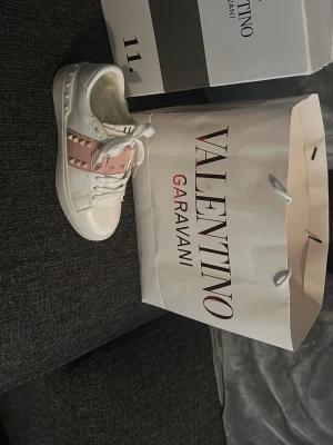 Valentino Garavani vita sneakers - Snygga vita sneakers från Valentino Garavani med rosa detaljer och nitar. Skorna har klassisk låg modell, vita skosnören och är tillverkade i läder. Perfekta för dig som vill sticka ut med exklusiva och trendiga skor.