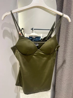 Olivgrön crop top med tunna band - Snygg olivgrön crop top med tunna axelband och markerade kupor. Toppen har en figurnära passform och är tillverkad i ett stretchigt, mjukt material som sitter skönt mot kroppen. Perfekt att styla med jeans eller kjol för en trendig look.