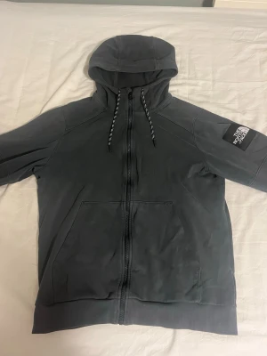Mörkgrå hoodie från The North Face - Snygg mörkgrå hoodie från The North Face med dragkedja framtill, huva med snören och två stora fickor. Logga på vänster ärm. Perfekt för chill dagar eller när du vill ha en clean streetwear-look.