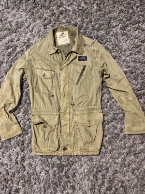 Morris olivgrön fieldjacket  - Snygg olivgrön vindjacka med fyra stora fickor framtill, dragkedja och knappar. Jackan har en klassisk krage och justerbar midja med snöre. Materialet är tunt och passar perfekt för blåsiga dagar. Detaljer som axelklaffar och patch på bröstet ger en cool look.