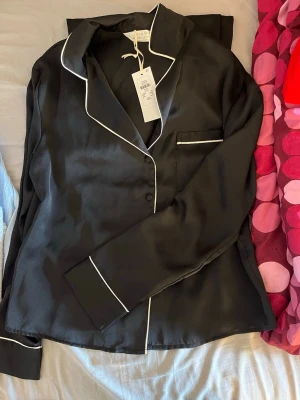 Svart satinpyjamas med vita detaljer - Stilren svart pyjamas i satin med vita kontrastkanter. Setet består av en långärmad skjorta med krage och bröstficka samt matchande byxor med resårmidja och vit dragsko. Perfekt för mysiga kvällar hemma.