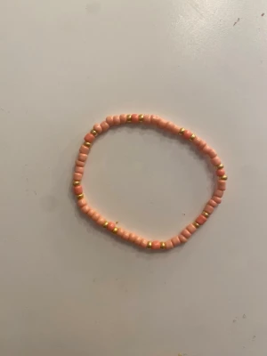 Rosa pärlarmband med gulddetaljer - Sött armband med små rosa pärlor och diskreta guldfärgade detaljer. Armbandet är elastiskt och passar de flesta handleder. Perfekt för att addera lite färg till din outfit.