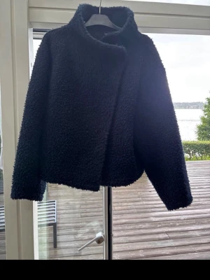 Svart teddyjacka med bred krage - Mysig svart teddyjacka med bred krage och avslappnad passform. Jackan har en fluffig struktur och stängs framtill. Perfekt för kyliga dagar och enkel att styla till jeans eller kjol. Använd fåtal gånger. 