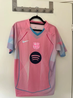 Rosa FC Barcelona matchtröja Nike - Säljer en rosa FC Barcelona matchtröja från Nike med Spotify-logga på bröstet. Tröjan har korta ärmar, V-ringning med blå kant och vita grafiska linjer framtill. Materialet är lätt och andas, perfekt för träning eller match.
