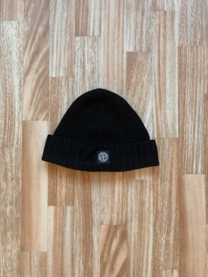 Svart ribbstickad Stone Island mössa - Säljer en svart ribbstickad stone island mössa/beanie med uppvikt kant och en rund patch framtill. Mössan har en enkel och stilren design, men man kan se några defekter. No size men passar bäst på tonåringar