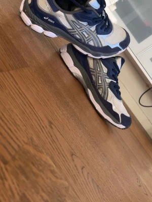 Blå och vita Asics sneakers - Snygga sneakers från Asics i blått och vitt med grå detaljer. Skorna har meshpaneler för bra ventilation och klassisk Asics-logga på sidan. Yttersulan är vit med grå inslag och ger bra grepp. Perfekta för dig som gillar sportig och stilren look.