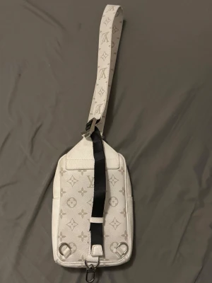 Vit axelväska med monogram - Snygg vit axelväska i rektangulär form med bred axelrem och klassiskt monogrammönster. Väskan har silverfärgade detaljer, dragkedja och en praktisk ytterficka. Materialet ser ut att vara tåligt canvasliknande tyg.