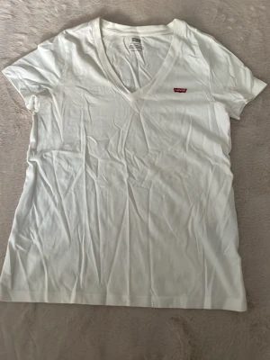 Vit v-ringad t-shirt från Levi's - Klassisk vit t-shirt från Levi's med v-ringning och liten röd logotyp på bröstet. T-shirten har korta ärmar och är gjord i mjuk bomull, perfekt för en enkel och clean look. Tröjan är xxs men passar dig som har storlek xs och s. 